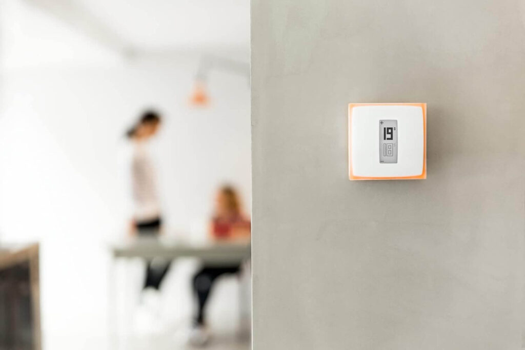 "Thermostat intelligent monté sur un mur avec deux personnes en arrière-plan."