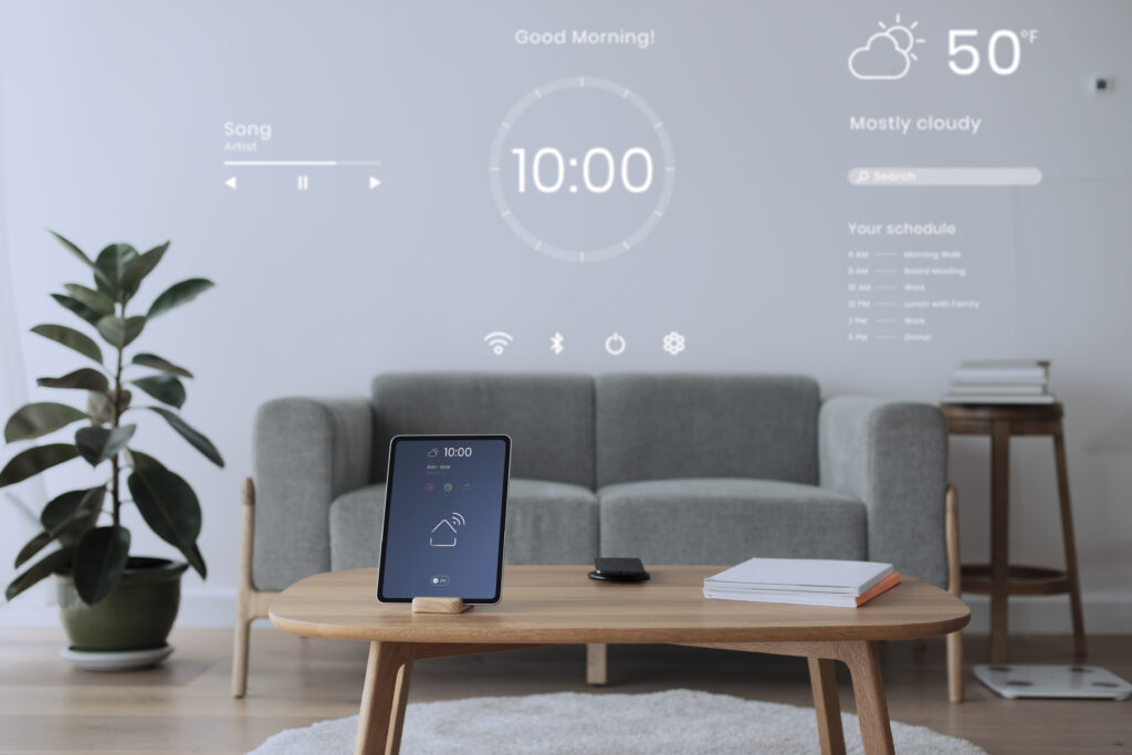 Tablette affichant un contrôleur de maison connectée sur une table en bois, avec un canapé gris et des éléments de projection d'informations sur le mur (horloge, météo, agenda)